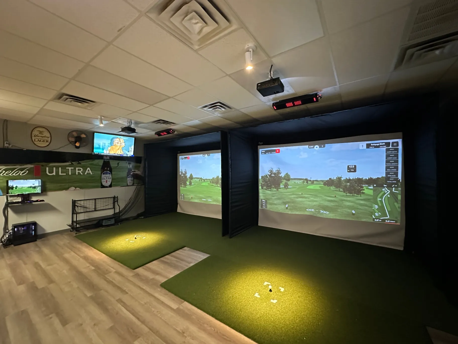 A modern golf simulator room with two large projection screens, a putting green, and a TV displaying sports, featuring a casual atmosphere
