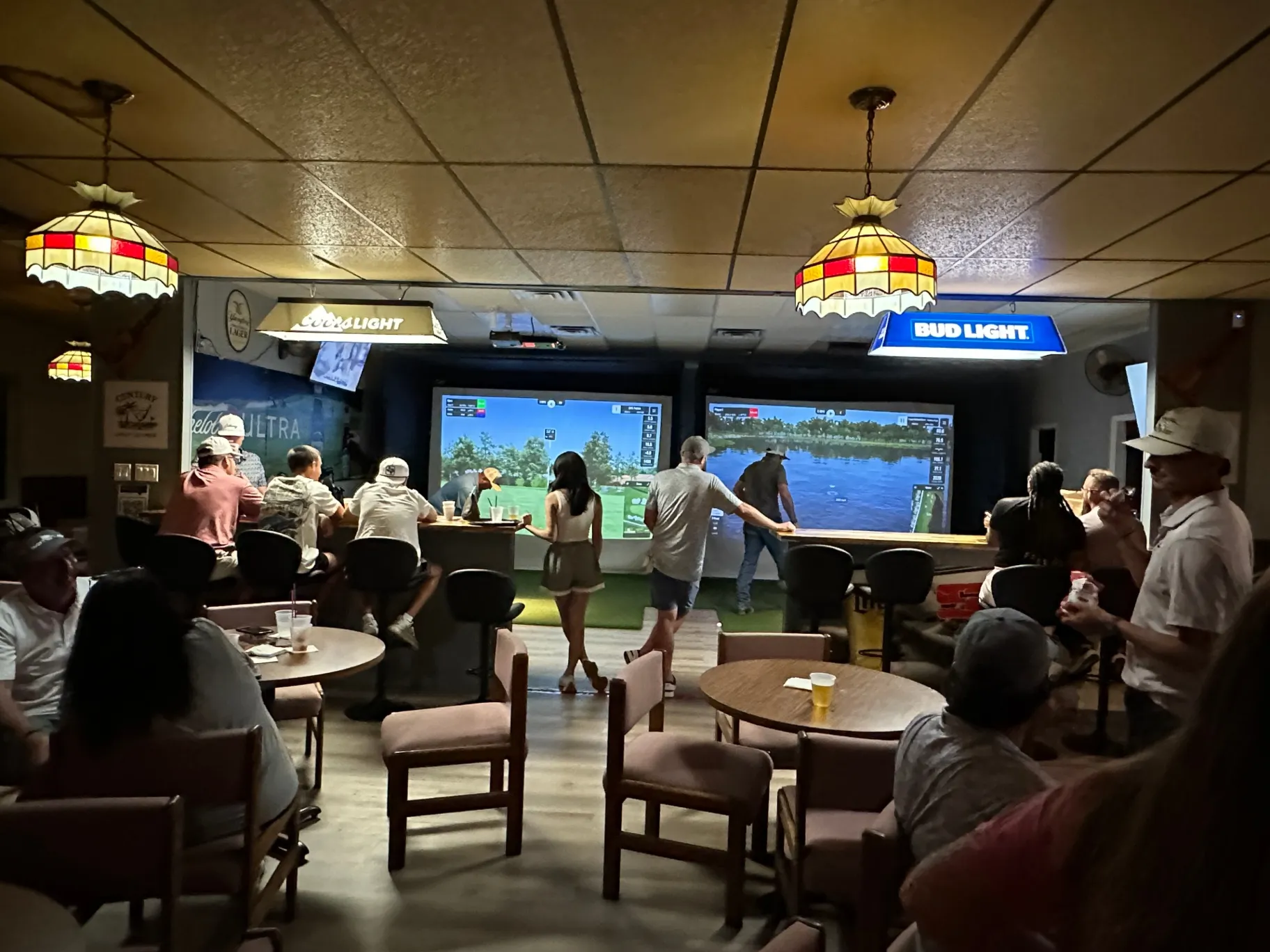 Group of people enjoying golf simulations in a lively bar setting with vintage lamps and beer advertisements