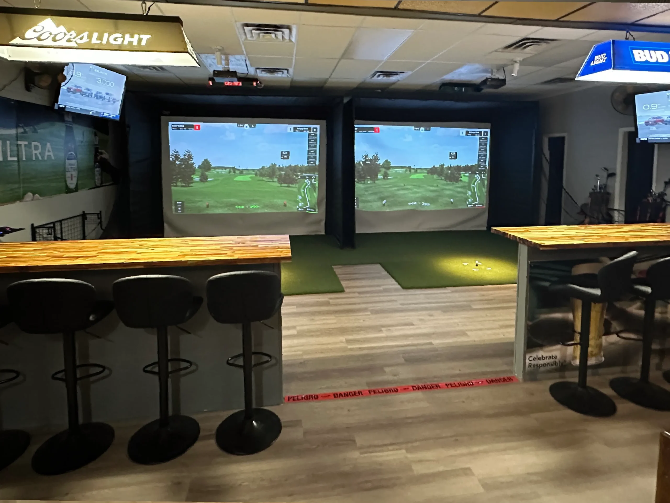 Indoor golfing facility featuring two projection screens, a wooden bar, and seating, with screens displaying golf courses and monitors